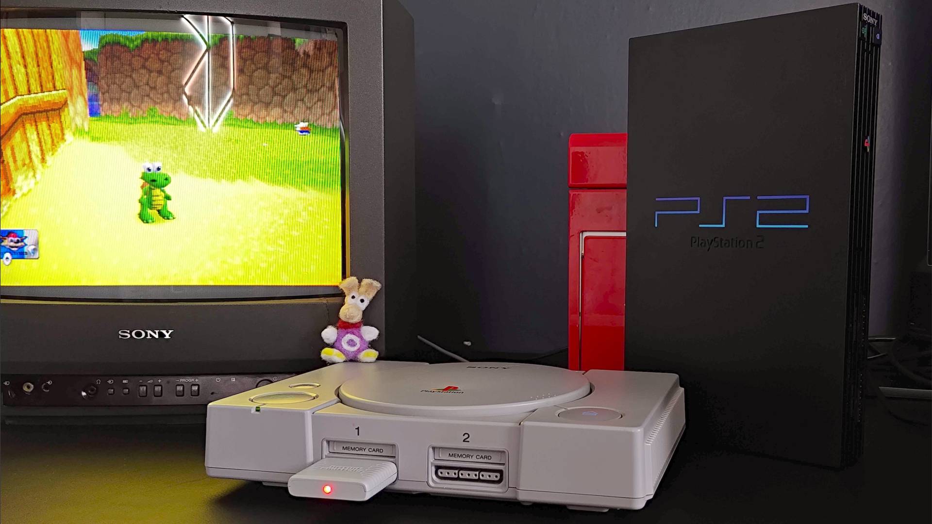 PS1 on black desk next to Sony Trinitron CRT TV with Croc 2 gameplay on screen and PS2 standing vertical on right.