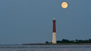 Large, bright full moon to the upper right of the image above a red and white lighthouse with water in the foreground. 