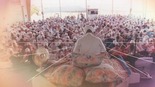 A man dressed in all white and a turban (Yogi Bhajan) sits on a pile of pillows in front of a crowd of followers dressed in all white, in 'Breath of Fire.'