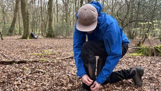 A man wearing a Revolution Race Aphex Pro is bending down adjust his trousers at the ankle, in a forest.