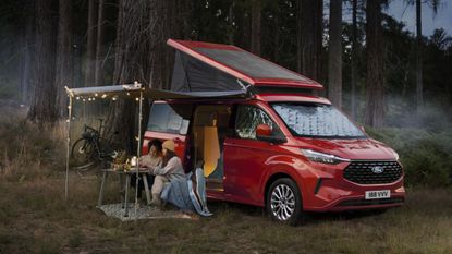 The Ford Nugget campervan in red, with people dining outside by some trees in a field