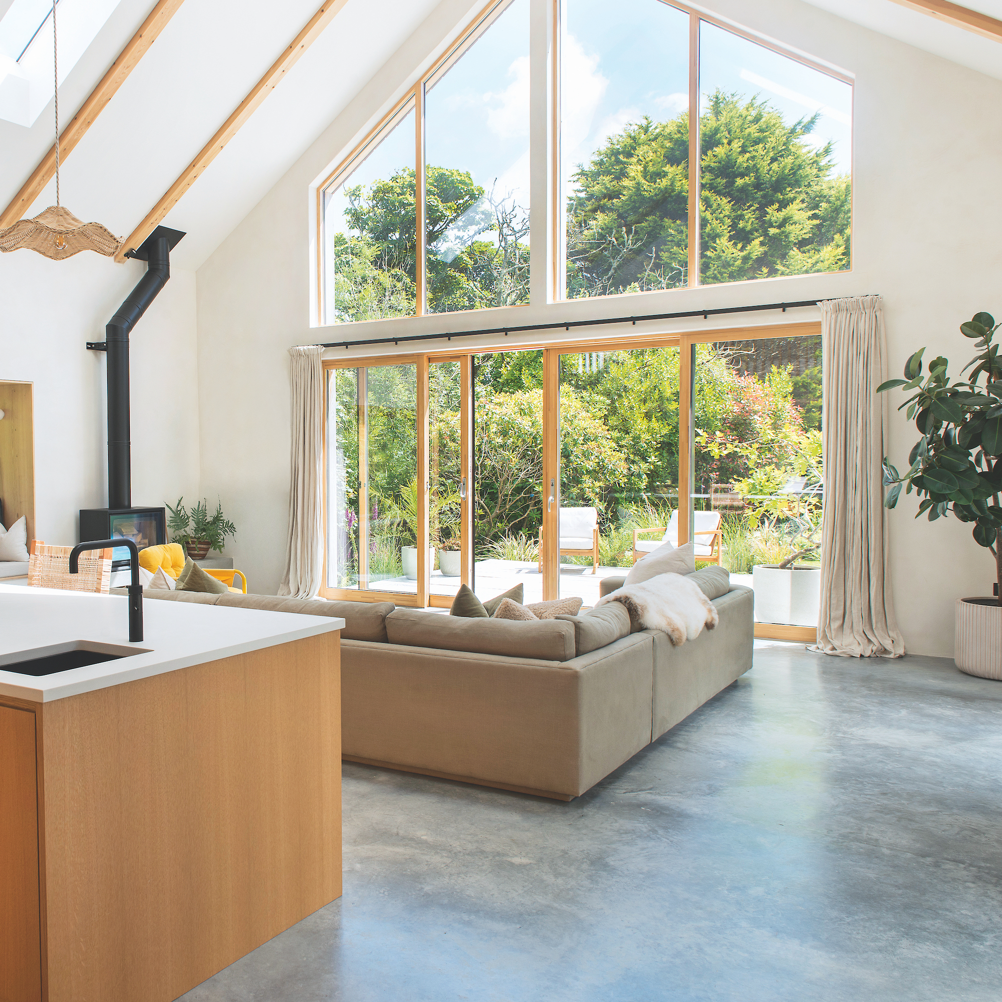 interior of self-build coastal house in cornwall featuring an open-plan living space with a kitchen and seating area, with grey corner sofa and large area of glazed wall with french doors and windows opening onto garden