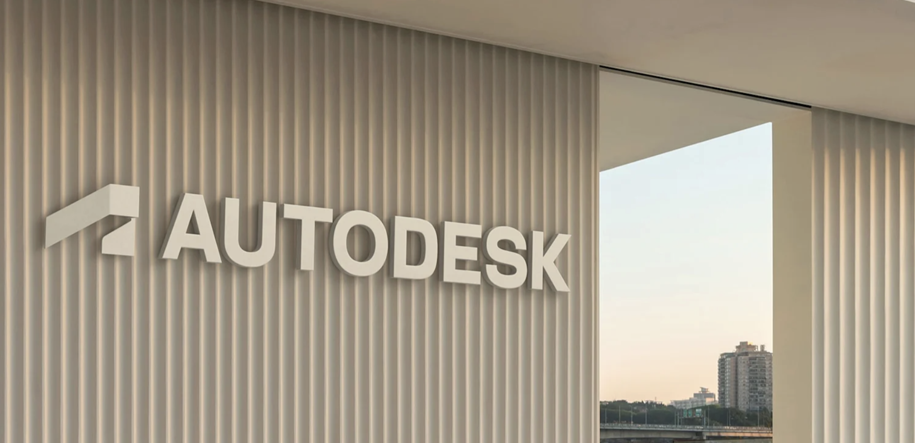Logotipo y denominación de Autodesk en 3D, blanco, limpio y minimalista, montado en una pared vertical de listones beige fuera de un edificio moderno.