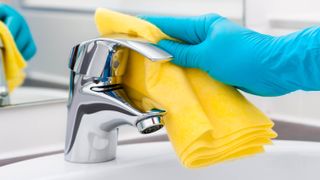 A faucet being cleaned with a microfiber cloth while wearing rubber gloves