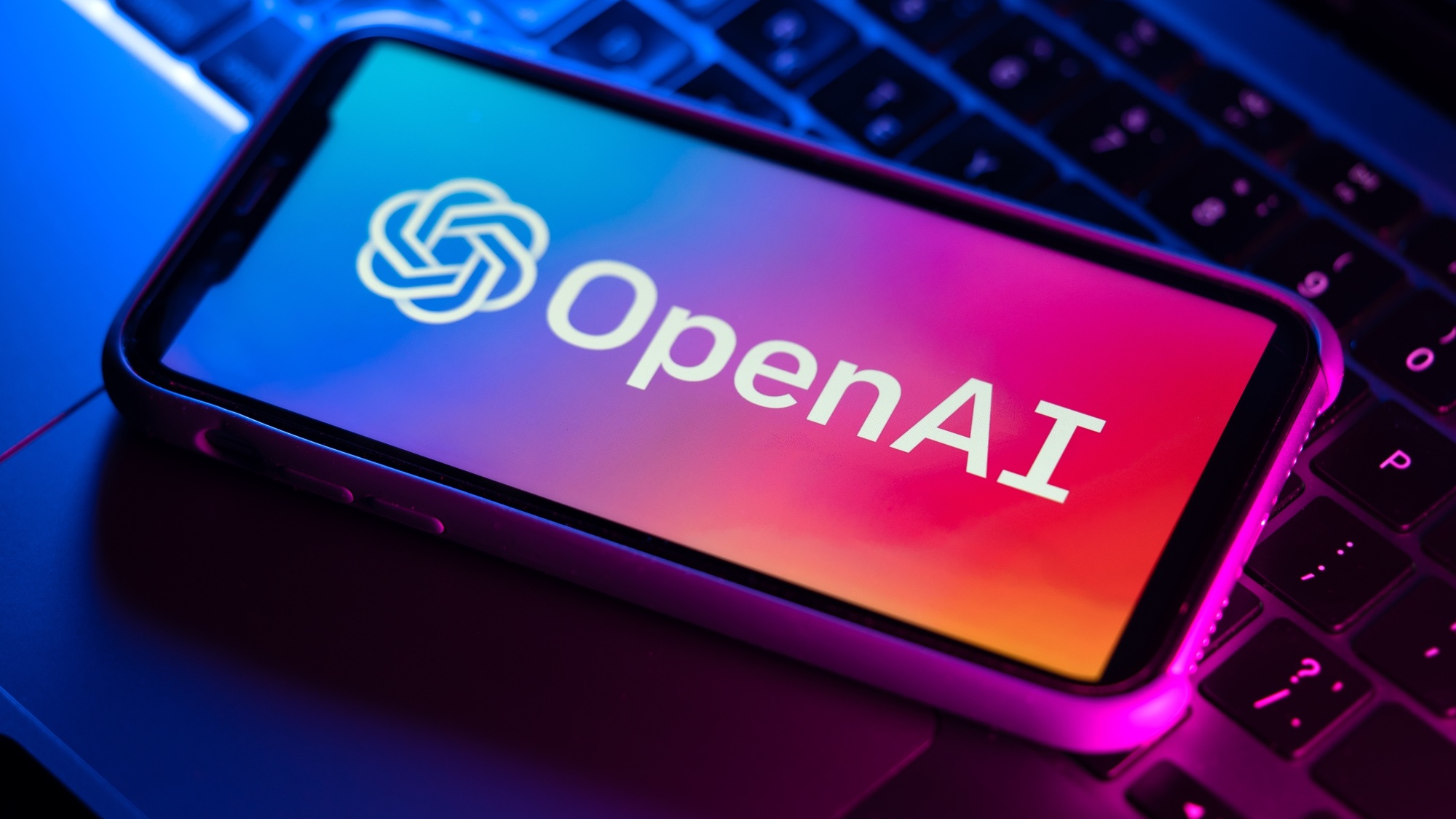 The OpenAI logo is on the phone above the laptop keyboard