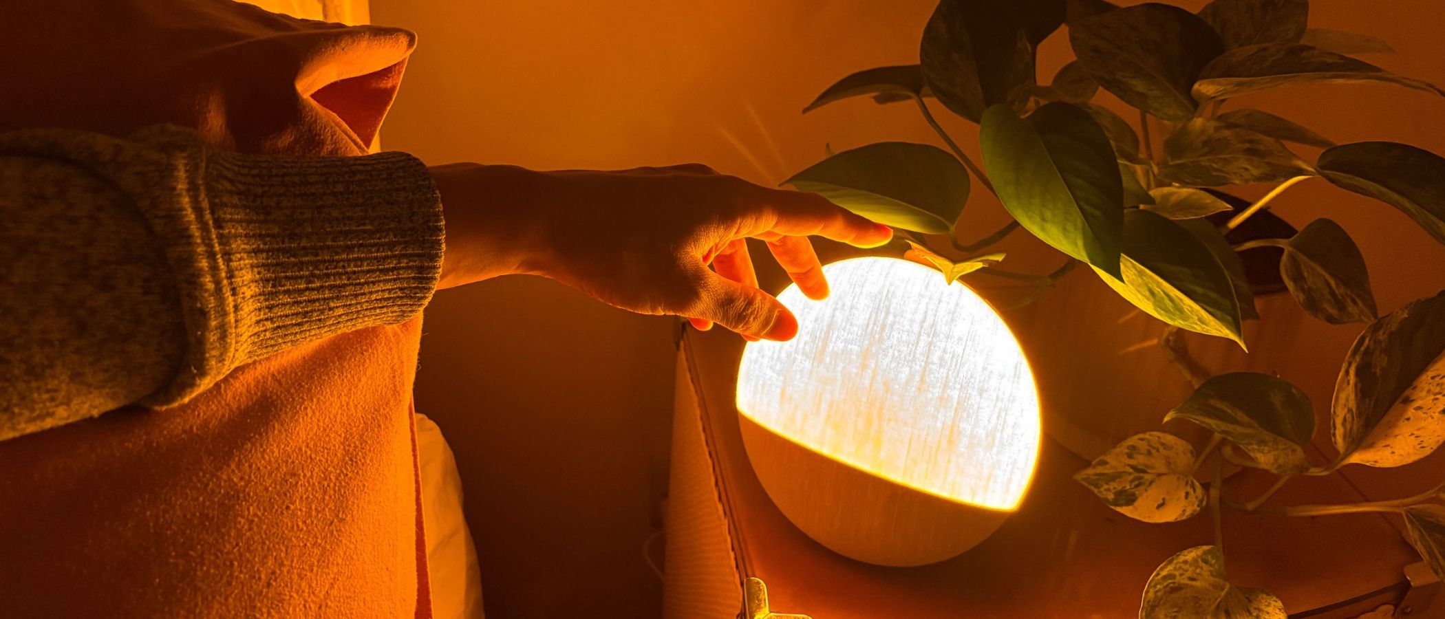 The image shows a circular night light placed on a nightstand emitting an orange glow against an orange and white pillow. From the left, a hand reaches over to it.