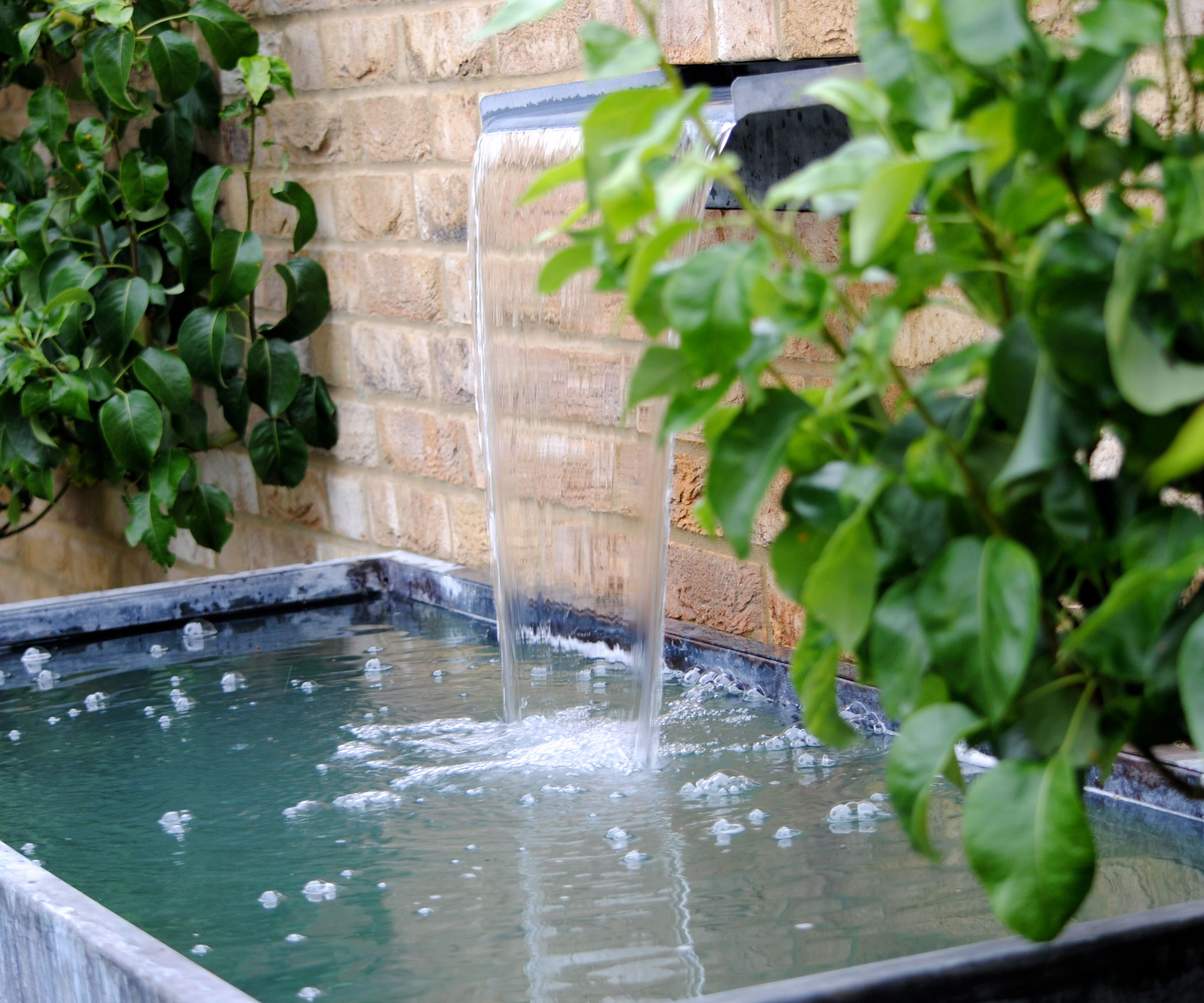 water feature with water blade and water tank, positioned against a brick wall with planting