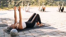 Woman leading Pilates class lying on back with arms in the air on yoga mat outdoors