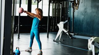 A woman in a sports vest, leggings and sneakers stands in a gym performing a kettlebell swing. Her body is upright, head facing forward and her arms are out in front holding the kettlebell. Behind her is a rowing machine and gym ropes.