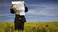 A businessman holds a map up in front of his face while standing in a field.