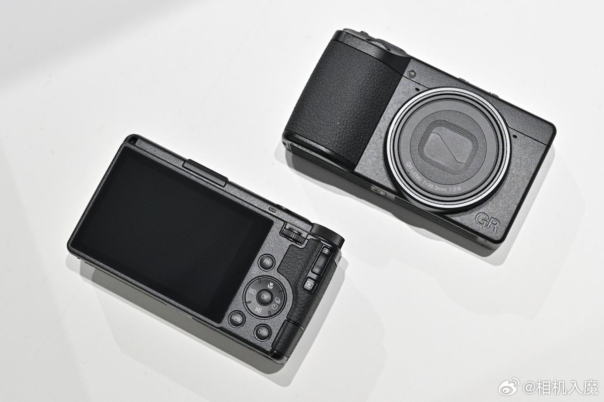 A Ricoh GR IV compact camera with a sleek design, showcasing its lens and rear LCD screen, placed on a light surface