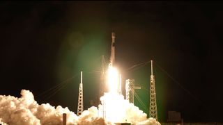 a white and black rocket lifts off into the night sky, its bright yellow-white plume lighting its launch pad below.