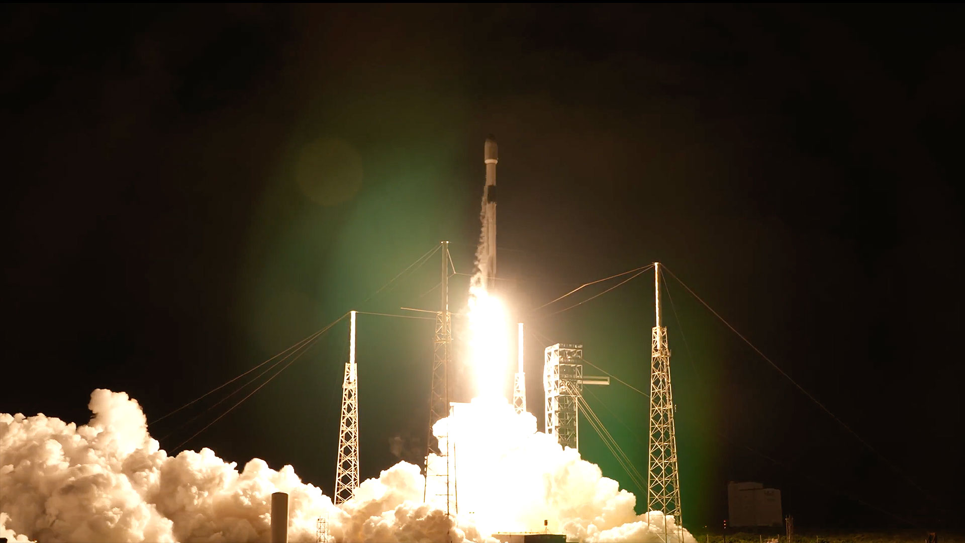 a white and black rocket lifts off into the night sky, its bright yellow-white plume lighting its launch pad below.