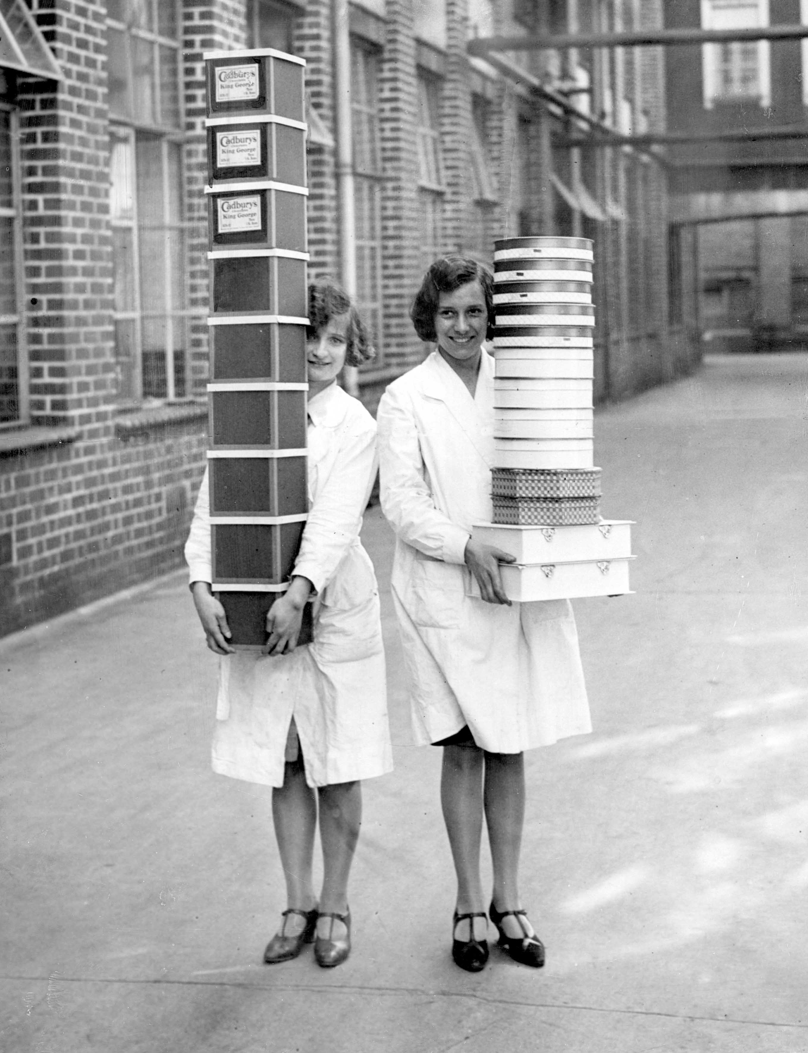Two smiling women in white overalls stand outside a brick factory building, one hugging a tall stack of Cadbury's chocolate boxes and the other carrying a pile of round and rectangular boxes.