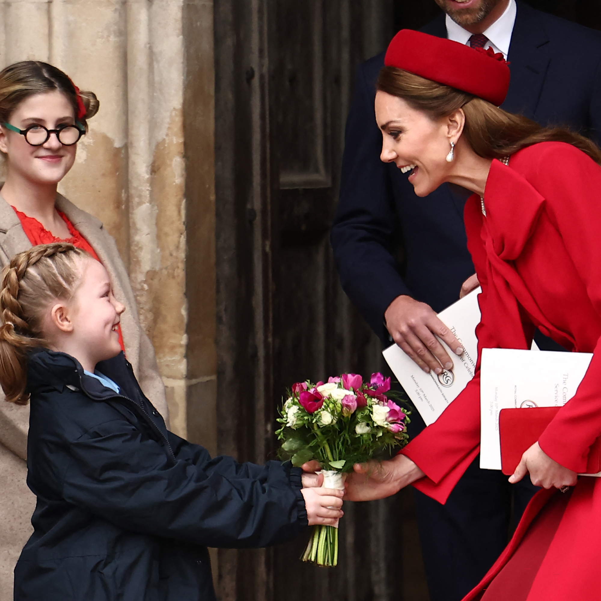 A little girl gives a bouquet to Kate Middleton dressed all in red