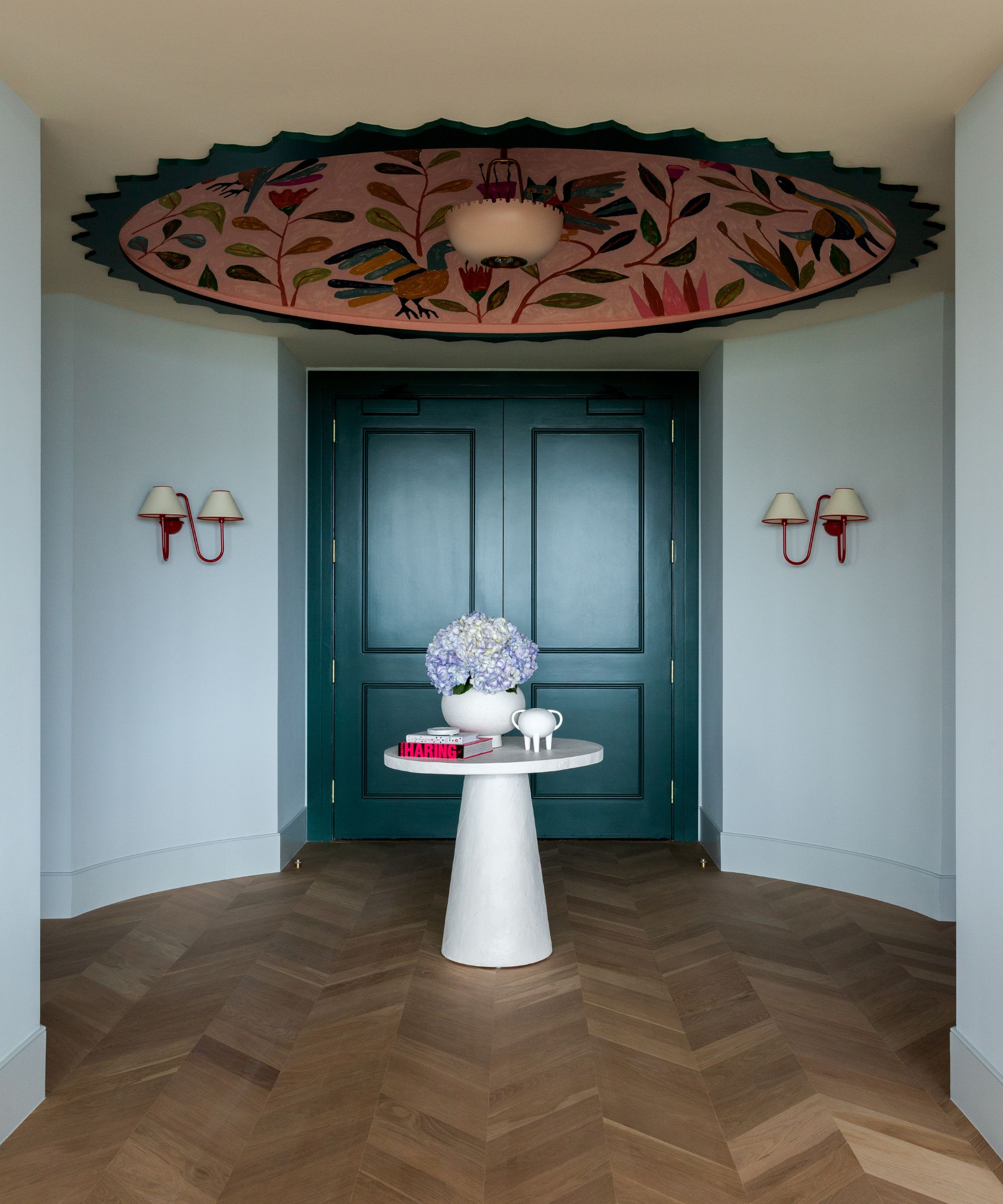 Pale blue entryway with dark teal double doors, two red sconces on walls, a large pink floral ceiling mural, a round, small white table in the centre with a vase and books, and parquet wooden floors