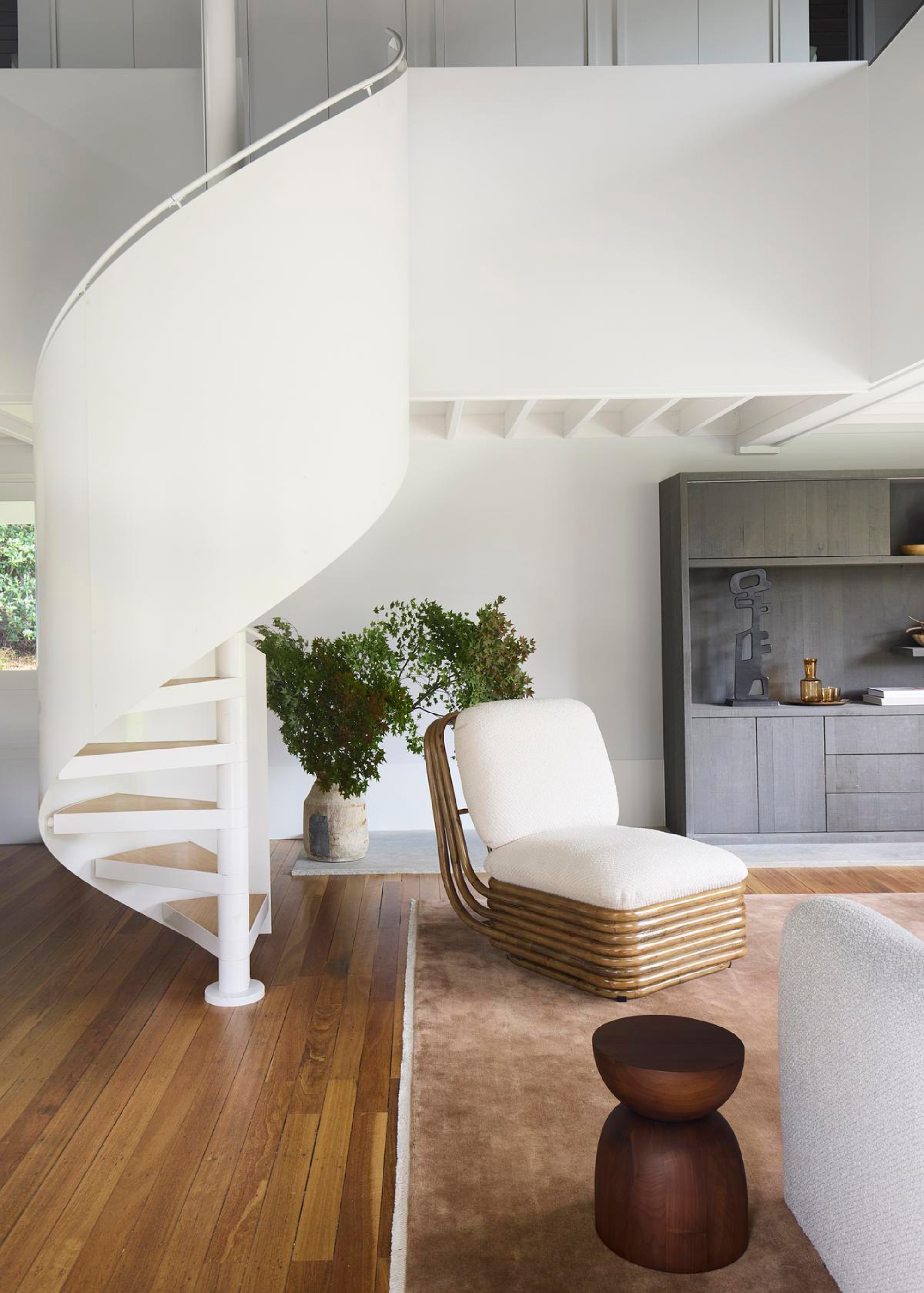 A living room with a white spiral staircase, a wooden stool, a vase of stems and a large rug
