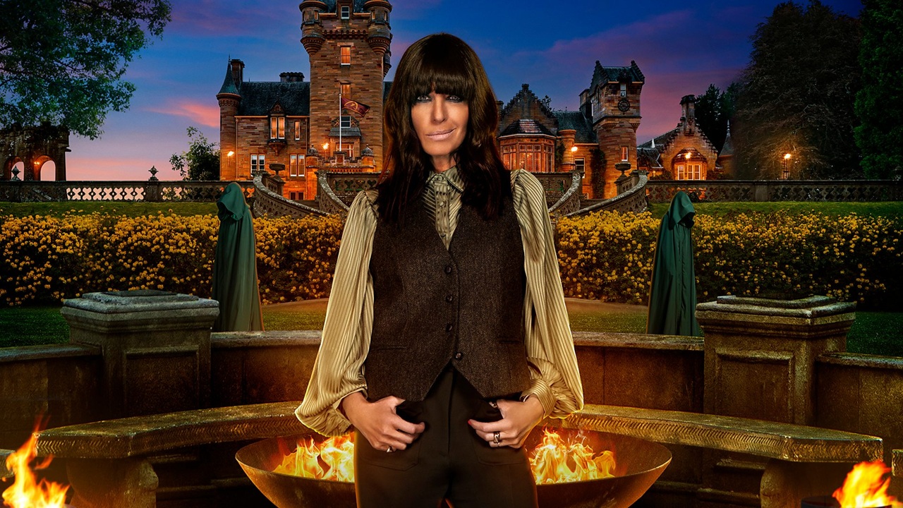 Host Claudia Winkleman stands in front of Ardross Castle at dusk, wearing a brown waistcoat and trousers and flanked by somber hooded figures and flames.