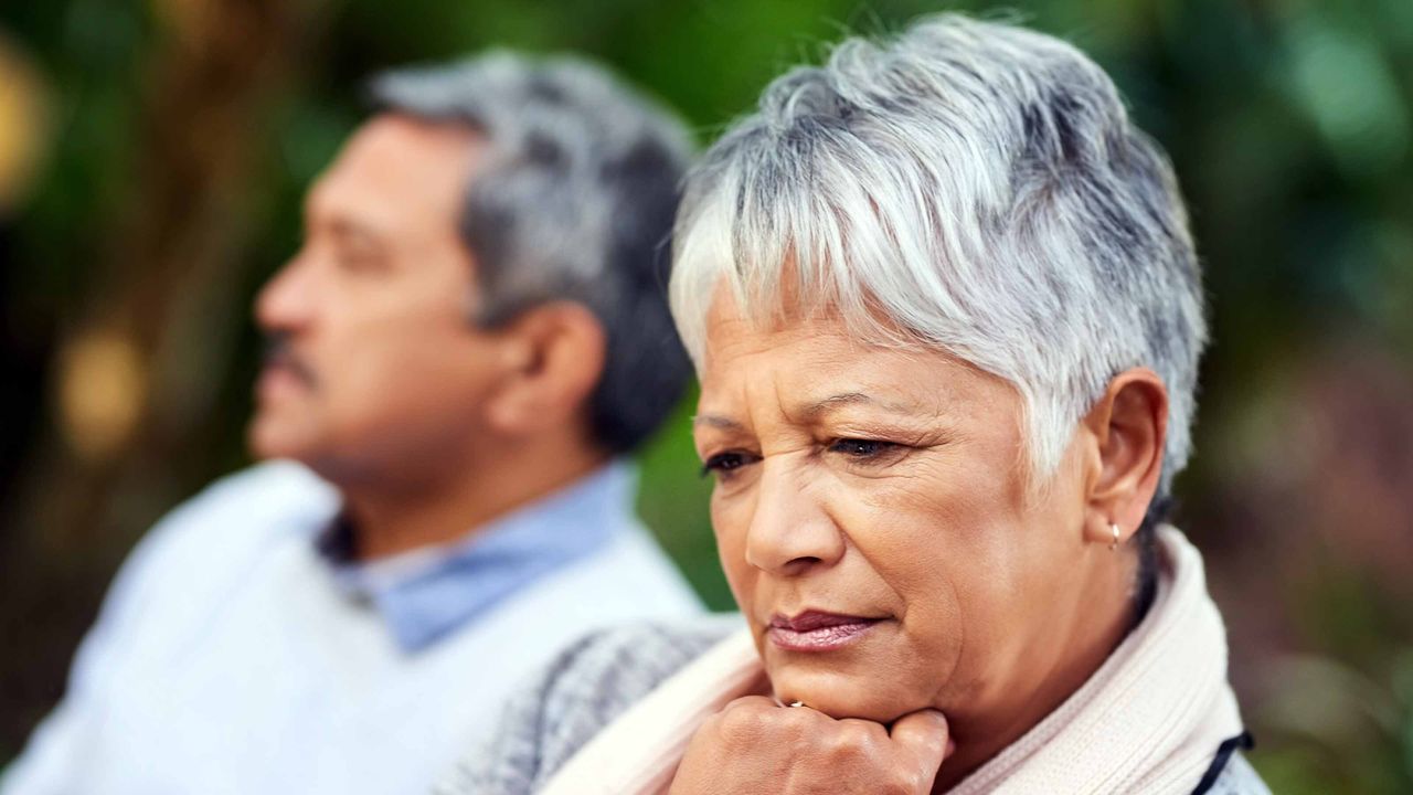 A woman with gray hair sitting outside in a sweater looks contemplative, looking in the opposite direction of a man sitting next to her.