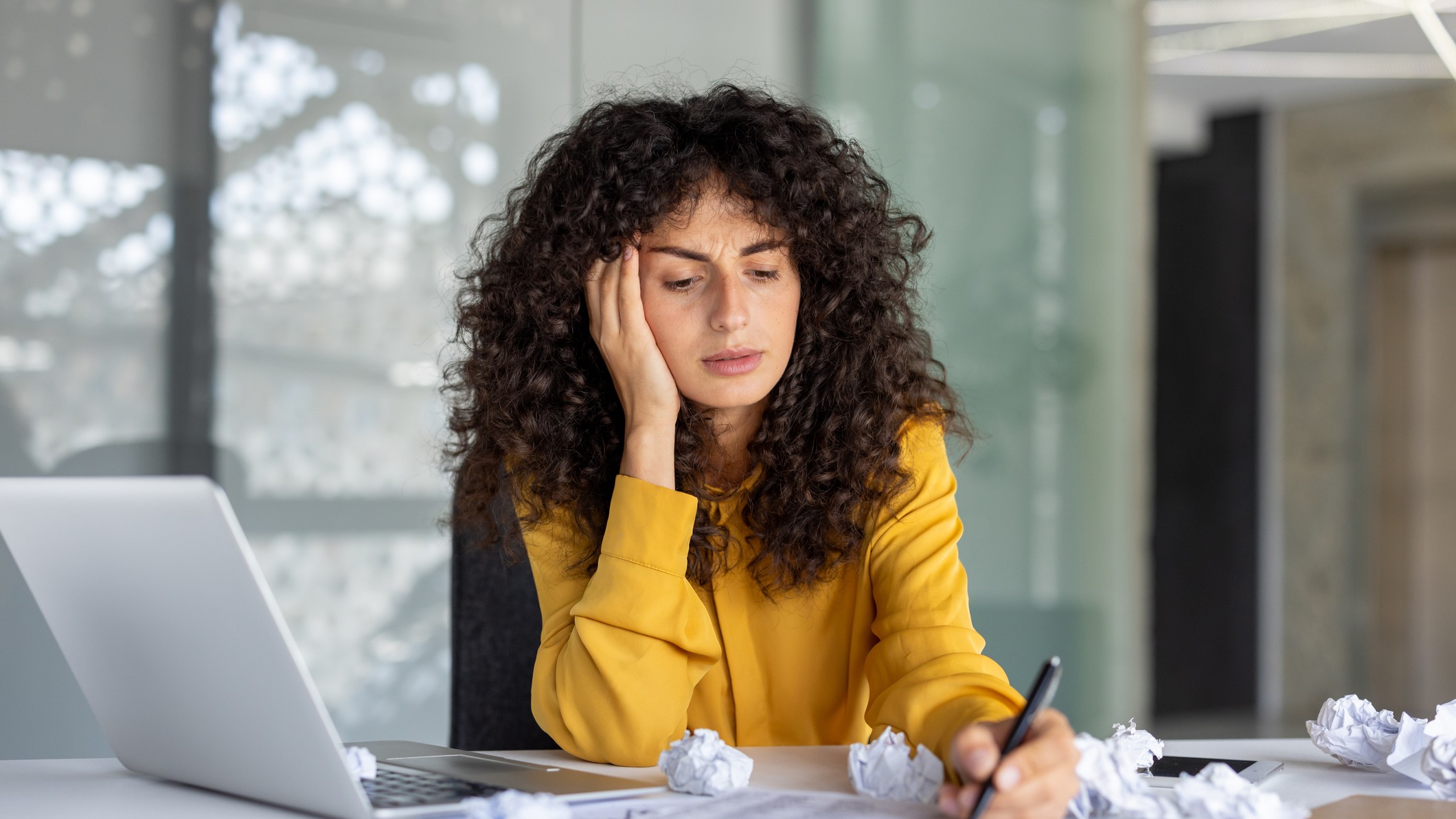 A woman attempts to write notes but looks confused, with crumbled pieces of paper around her. 