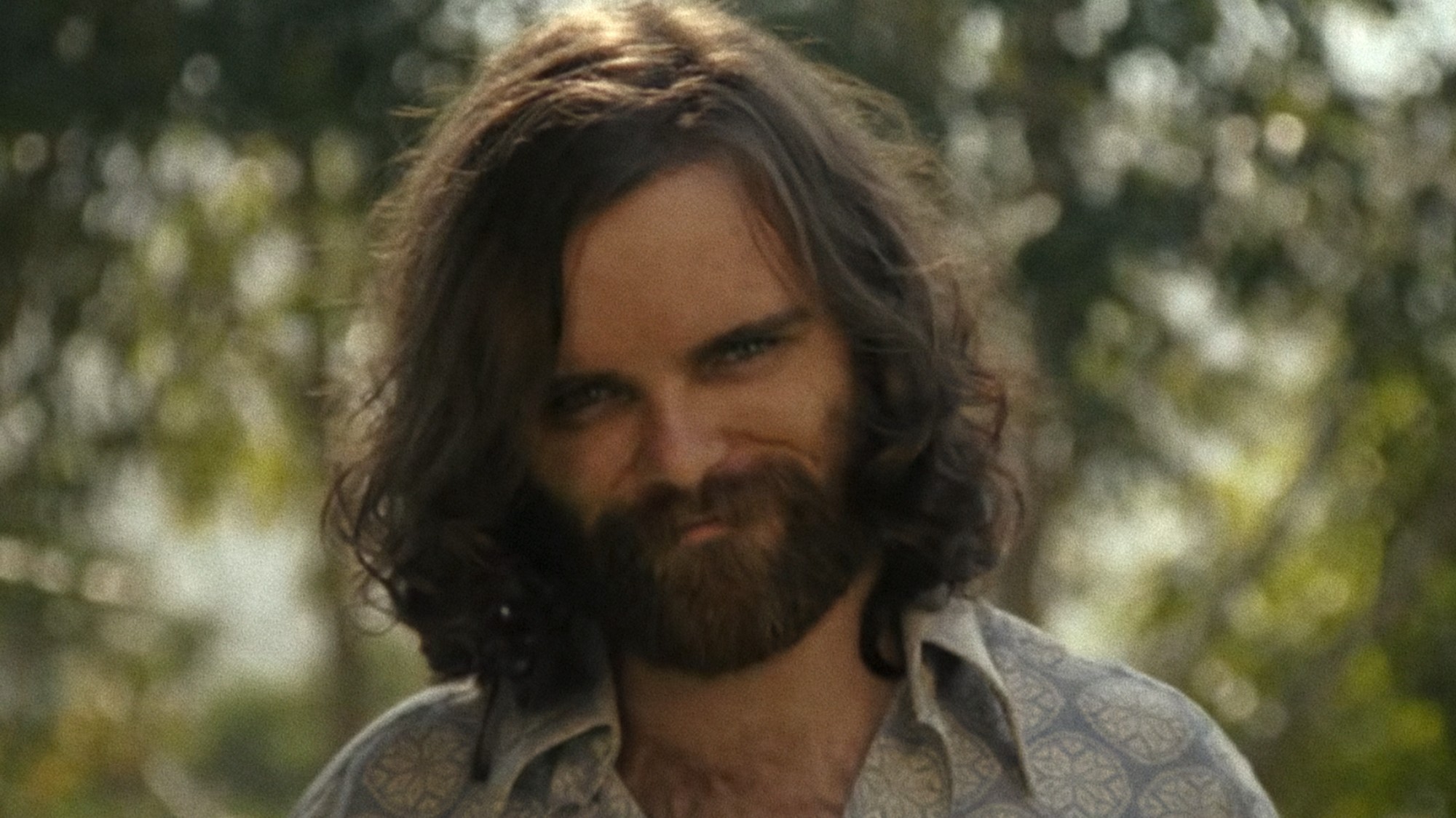 An actor playing Charles Manson with a beard and long hair, smiling and looking creepy