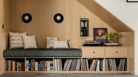 Image of a wooden reading and vinyl listening nook. There is a built-in bench seat with a gray cushion and built-in vinyl storage underneath. There is a record player on the side of the bench seat with a vinyl styled to show the cover art. 