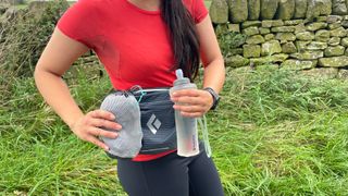 A woman wearing a Black Diamond Pursuit Waist Pack 6L holds a bag and soft water bottle in front of it to show how large it is.