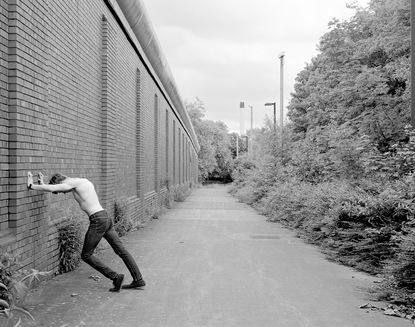 Black and white photo of man with no shirt standing on street with wall on one side and bushes on the other.