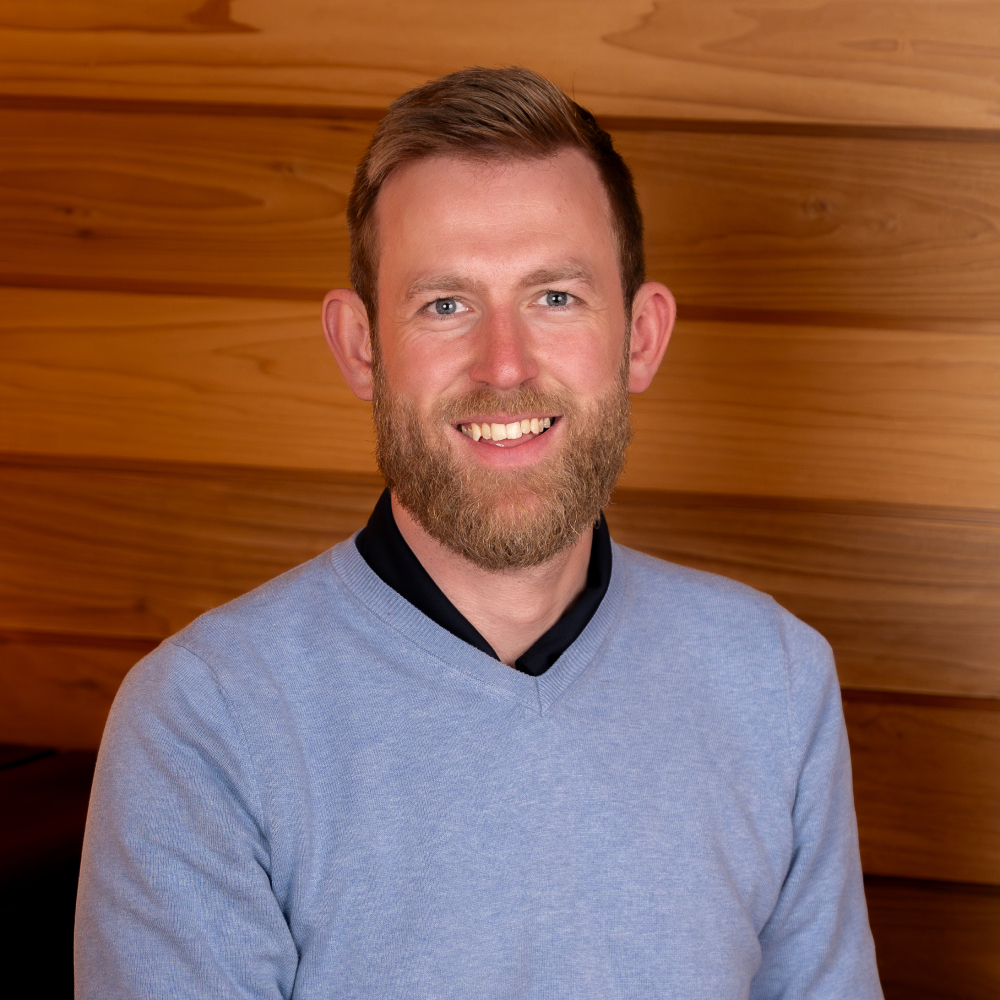 male with short hair and beard wearing jumper and shirt sat inside sauna