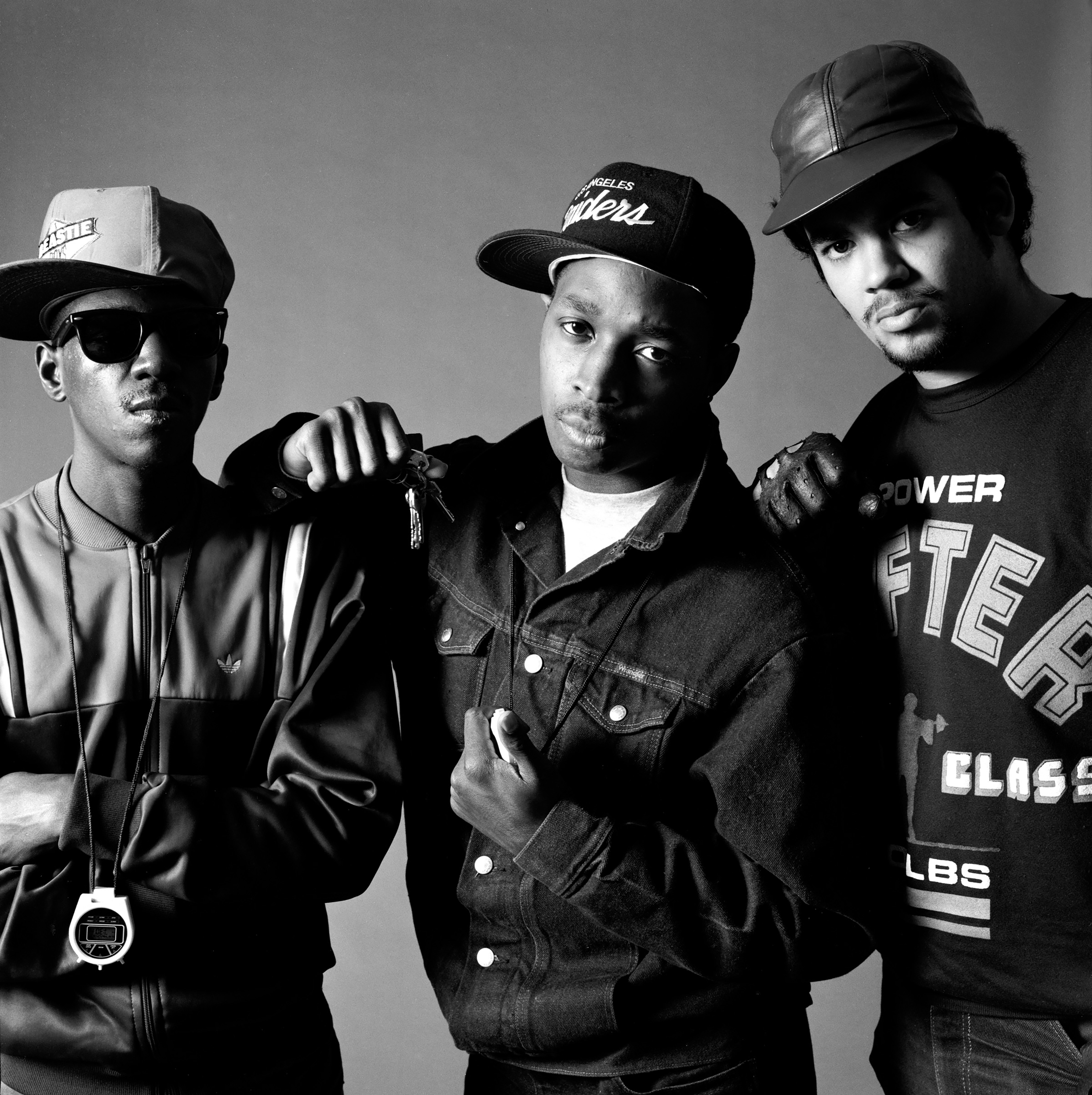 Black and white studio portrait of three members of Public Enemy wearing baseball caps and sportswear, posed close together against a grey background.