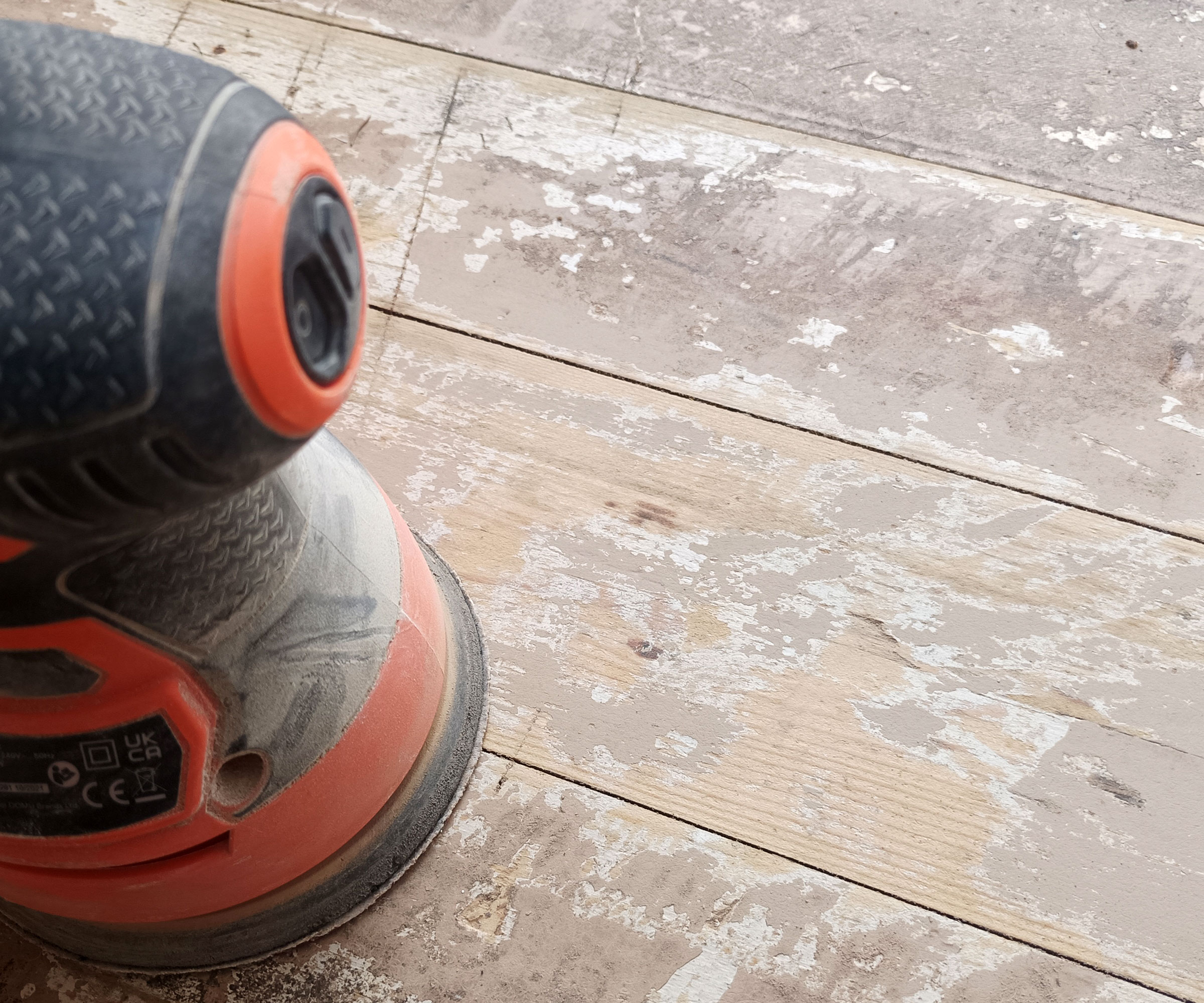 Orange and black orbital sander sat on a painted wooden floor wth bare wood exposed