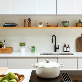 a kitchen island with an induction hob with a white casserole dish on top, a row of white kitchen cabinets, both wall hung and base cabinets with an inset kitchen sink and black tap