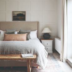 An off white-painted bedroom with a panelled feature wall behind the bed with an upholstered neutral-coloured headboard