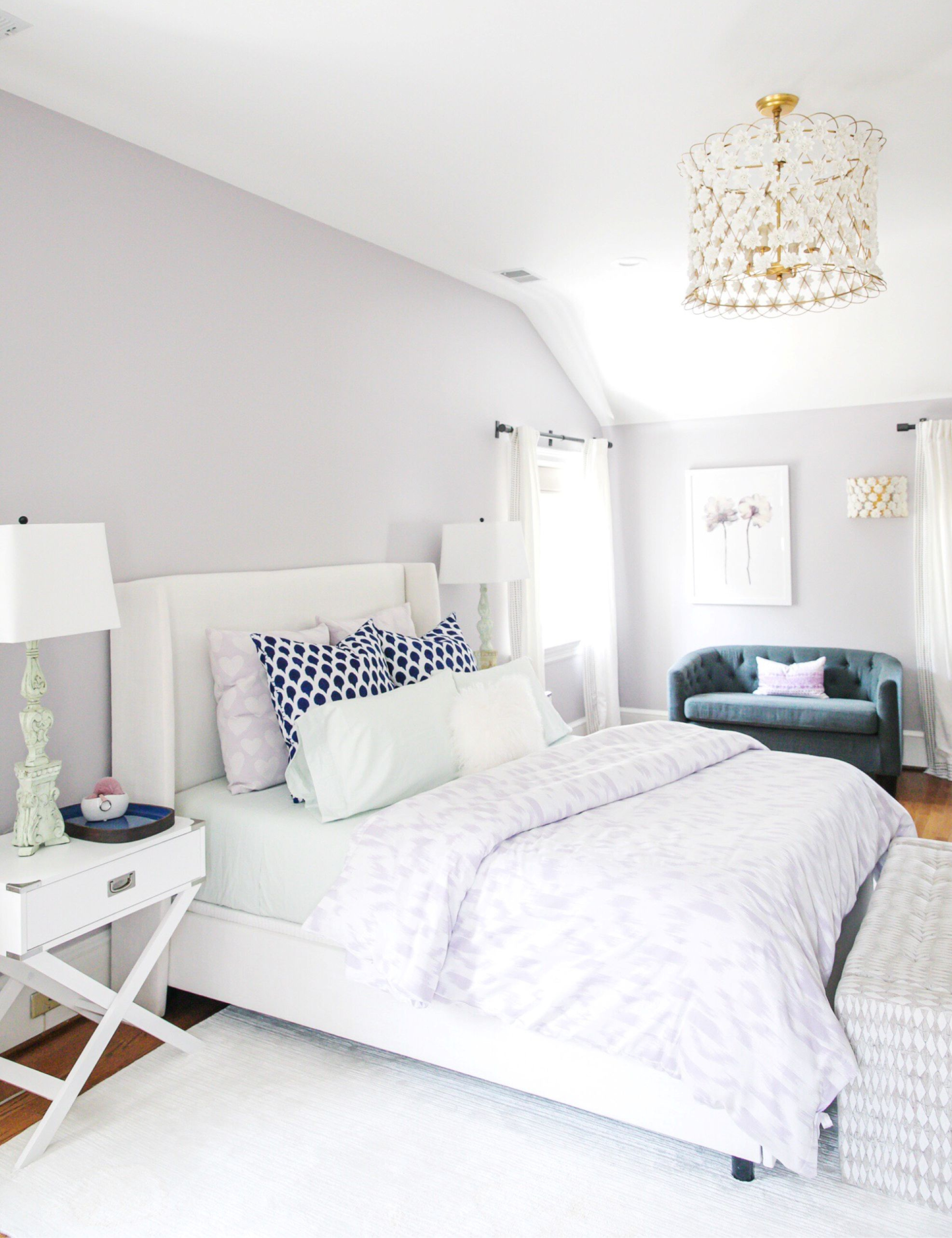 a bedroom with light gray walls, a white bed with purple sheets and blue pillows, a gold chandelier, and a white nightstand