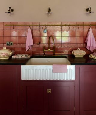 dark red kitchen with black countertop, fluted basin, glossy blush tiles, brass hanging rail, fluted shade wall lights, brass tap,