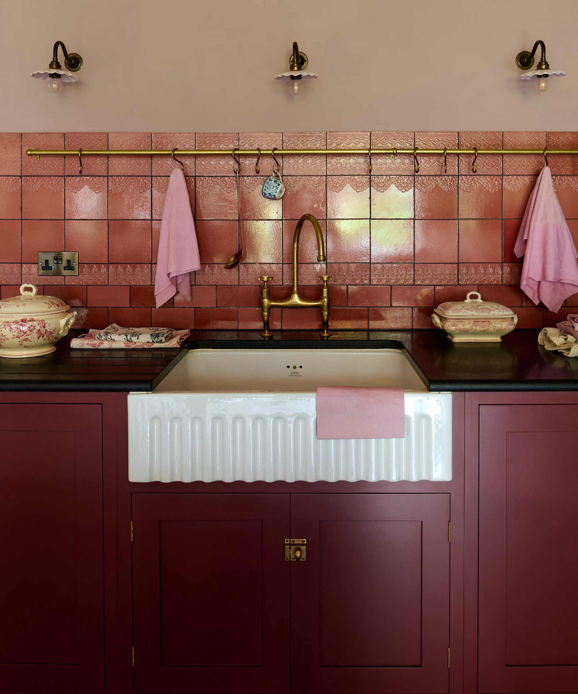 dark red kitchen with black countertop, fluted basin, glossy blush tiles, brass hanging rail, fluted shade wall lights, brass tap,