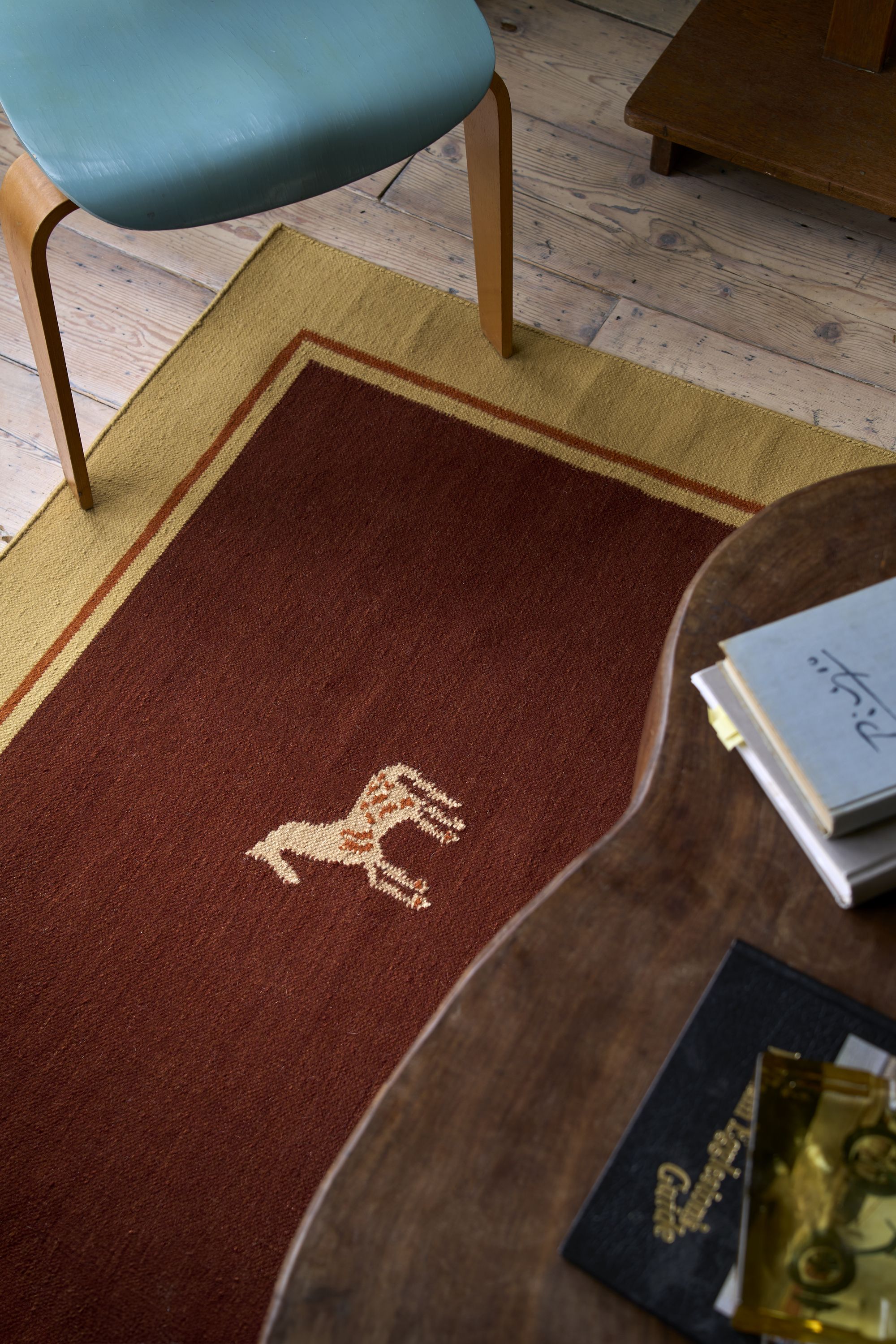 Close-up image of a red rug with a yellow horse on it and yellow trim. There is a teal accent chair and a wooden coffee table partially in view.