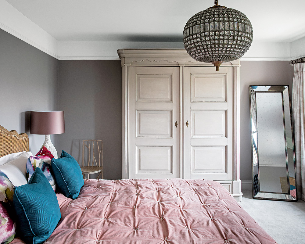 A bed with a pink throw and blue cushions next to a large, white wardrobe and a pink bedside lamp