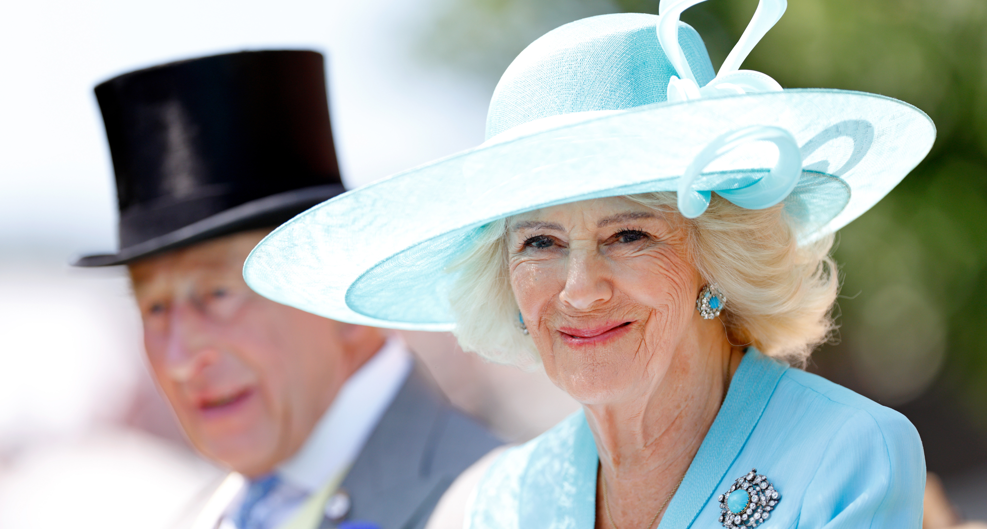 Queen Camilla in a turquoise hat and coat sitting next to King Charles
