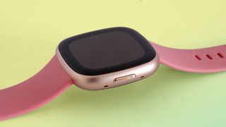 En Fitbit Versa 4 med ett rosa silikonband ligger på en ljusgul yta.