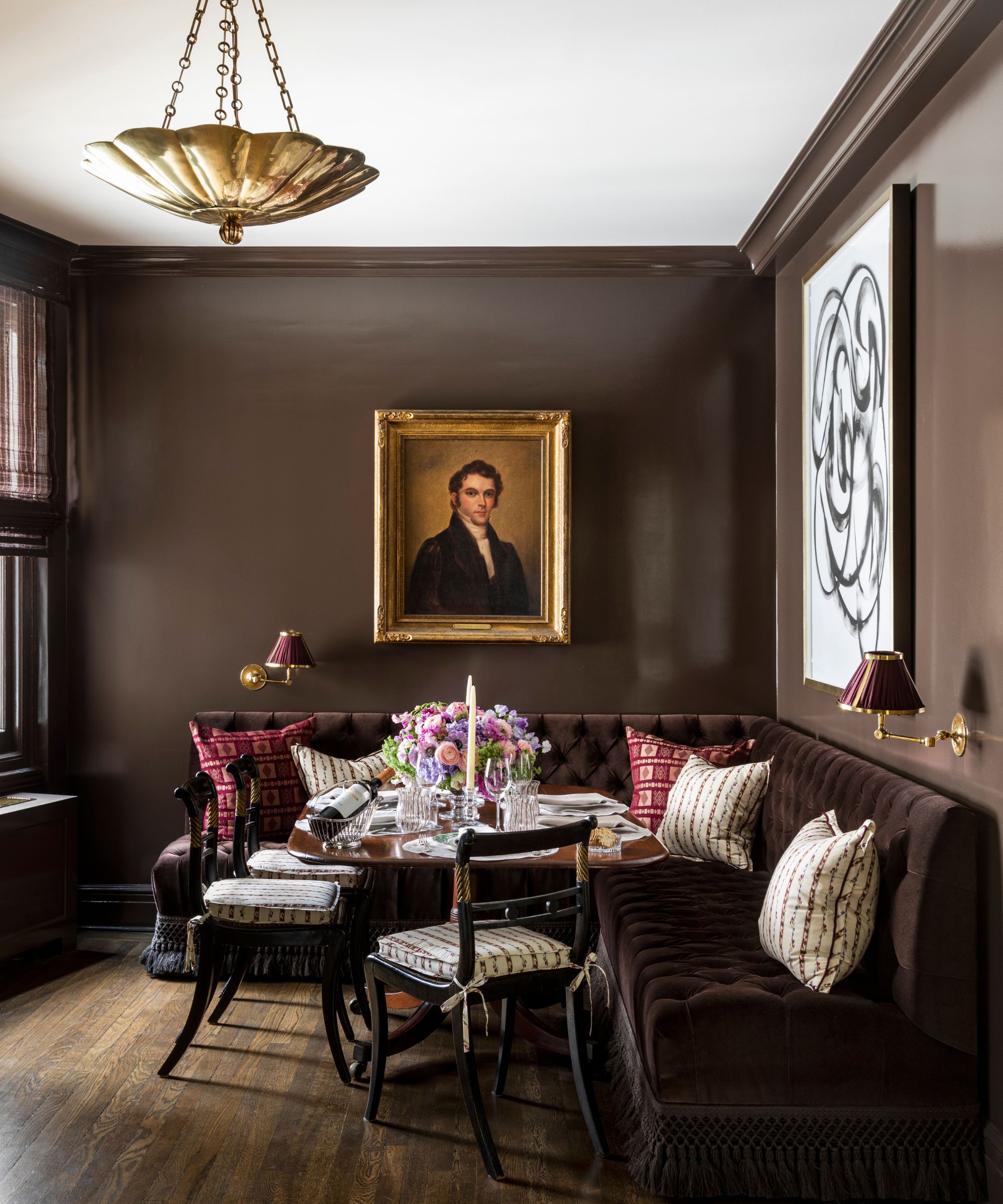 Dining room with dark brown walls, a gold flush-mount pendant, an antique portrait painting, a brown velvet banquette with patterned pillows, a wooden table, and three dark wood dining chairs.