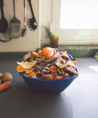 A dark blue plastic bowl filled with orange peels, banana skins, and onion peels, on a dark gray countertop with a white wall with kitchen utensils and a window behind it