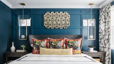 navy blue bedroom with white sheets, three patterned throw pillows and a bolster pillow on the bed, black bedside tables with ceramics, a sculptural mirror hanging over the bed and two pendant lights hanging over the side tables