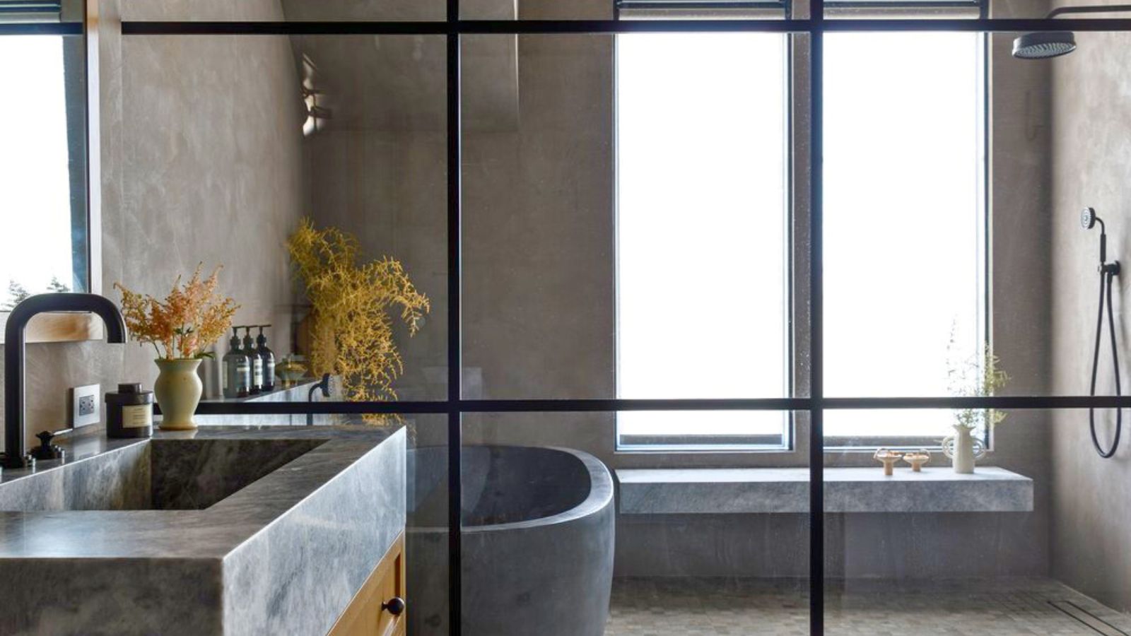Crittal glass shower in neutral stone bathroom. There is a shower seat and large frosted window visible, as well as gray stone bathtub and stone sink