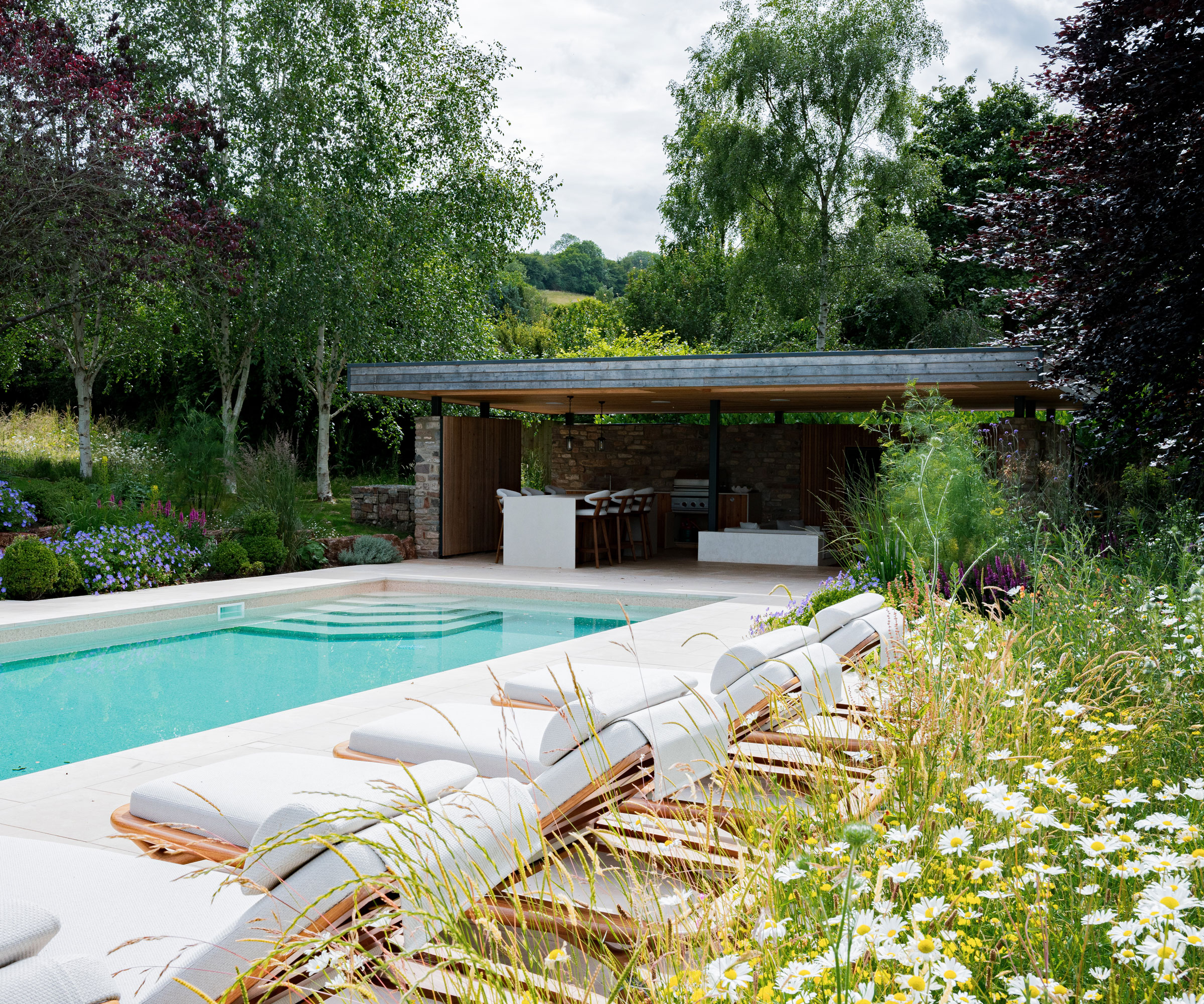 swimming pool and pool house with outdoor kitchen and dining area, sun loungers by the pool, planting and tall trees surrounding