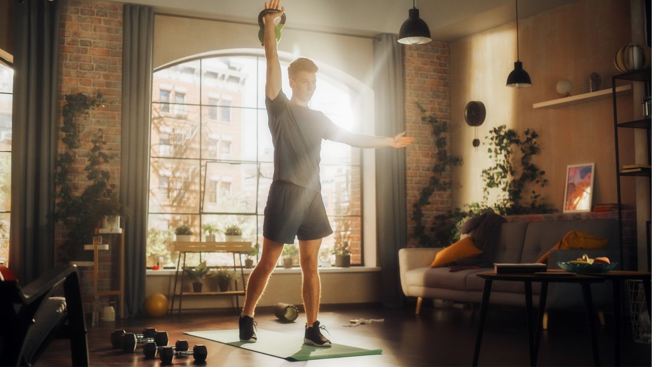 A man stands in a living room on an exercise mat, lifting a kettlebell above his head. He is in a split-legged stance, with one leg slightly in front of the other, the arm holding the kettlebell is pointed straight up while the other is out to the side. Behind him we see lots of green leafy plants, a couch, hanging lights and a big window.