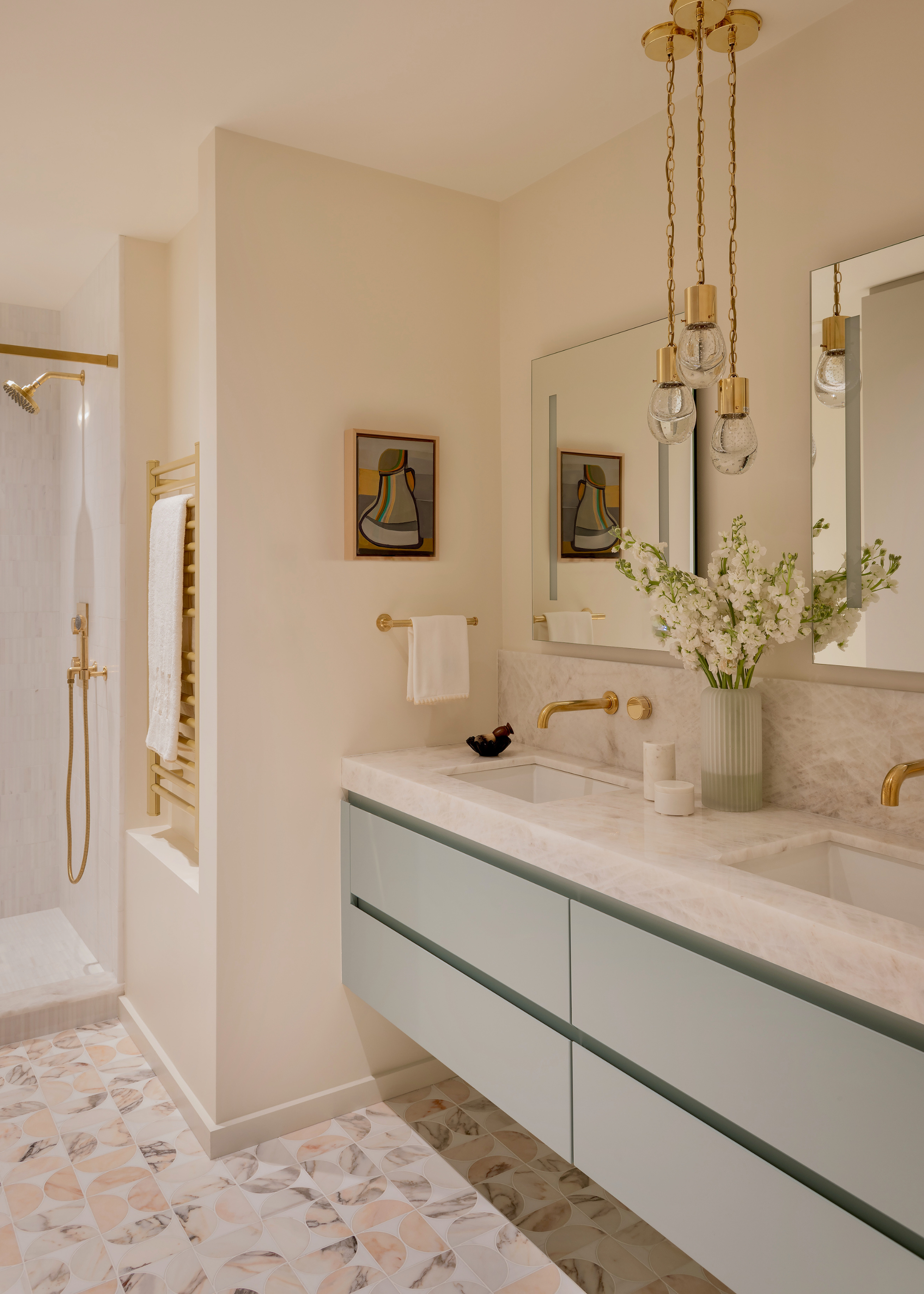 A modern bathroom with brass accents throughout, with a double vanity with light teal cabinets and marble countertops with two mirrors, a piece of art, a shower and towel rail in the background and a patterned tile flooring