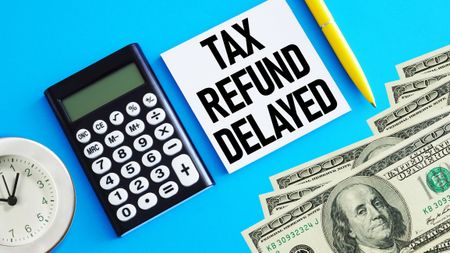 "tax refund delayed" printed on paper with money, a clock, a pen, and a calculator nearby