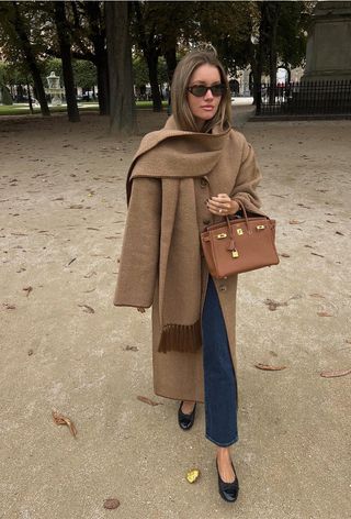 A French woman standing in a park wearing a tan coat with a matching cashmere scarf, a tan handbag, jeans, and black ballet flats.