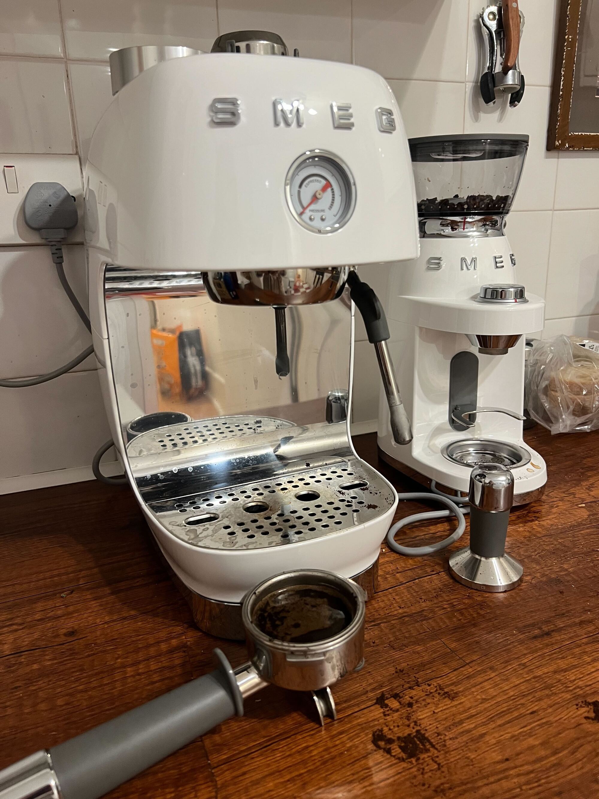 Image of a white, retro-style Smeg cold brew coffee machine that is slightly dirty on a wooden countertop with a portafiller that is filled with used coffee grinds.
